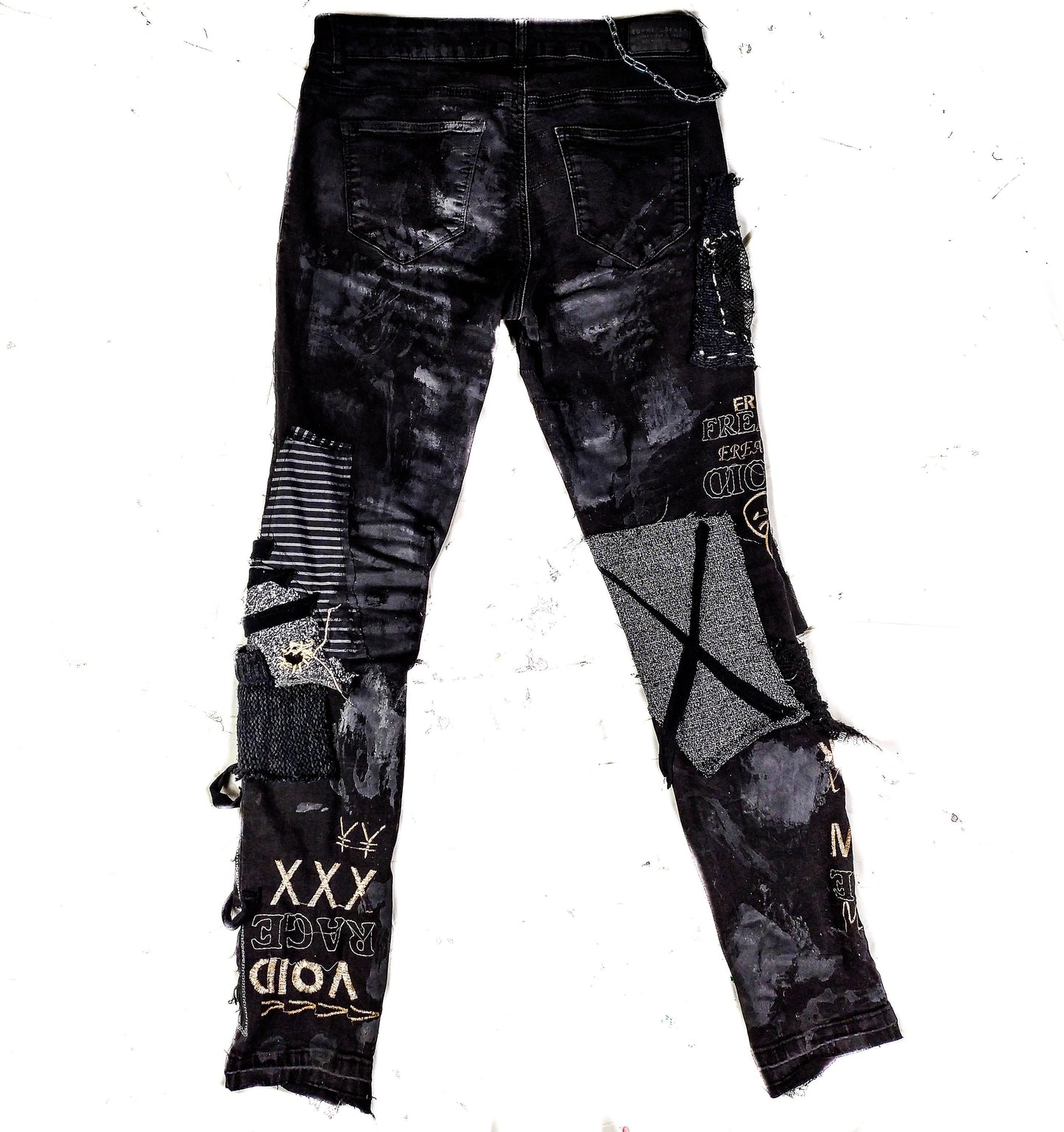 Flat laid black skinny pair with various abstract embroideries and words such as void walker and more riot with abstract symbols, black, white and grey patchwork, zippers, studs, splashes of gray and black paint and a chain attached to pocket.