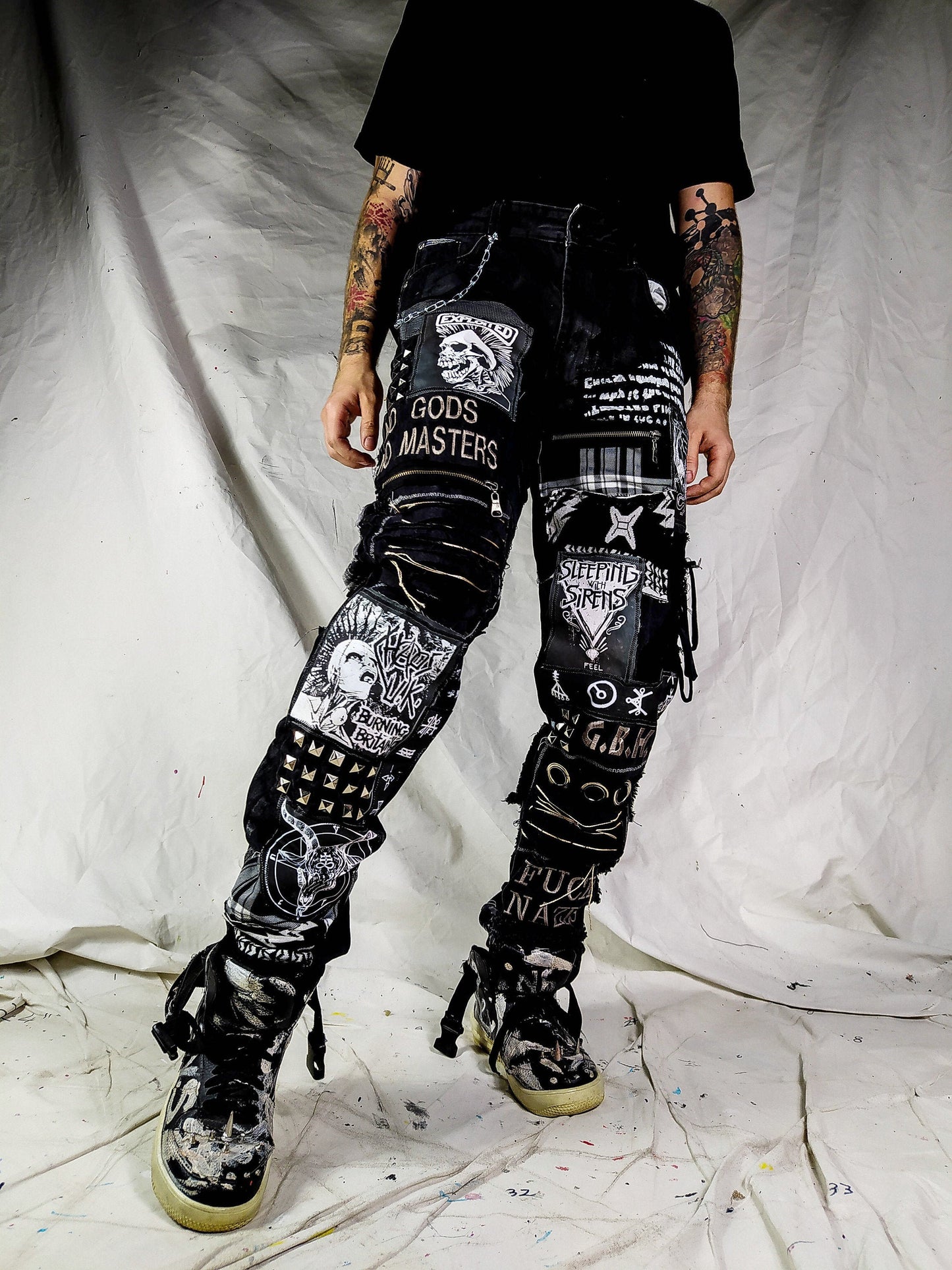 Model showing a black pair with embroideries such as no gods, no masters, prints with punk bands such as exploited, burning britain, sleeping with sirens along with studs, goat with horns, eyelets, laces, zippers, chain, abstract paint and patchwork.