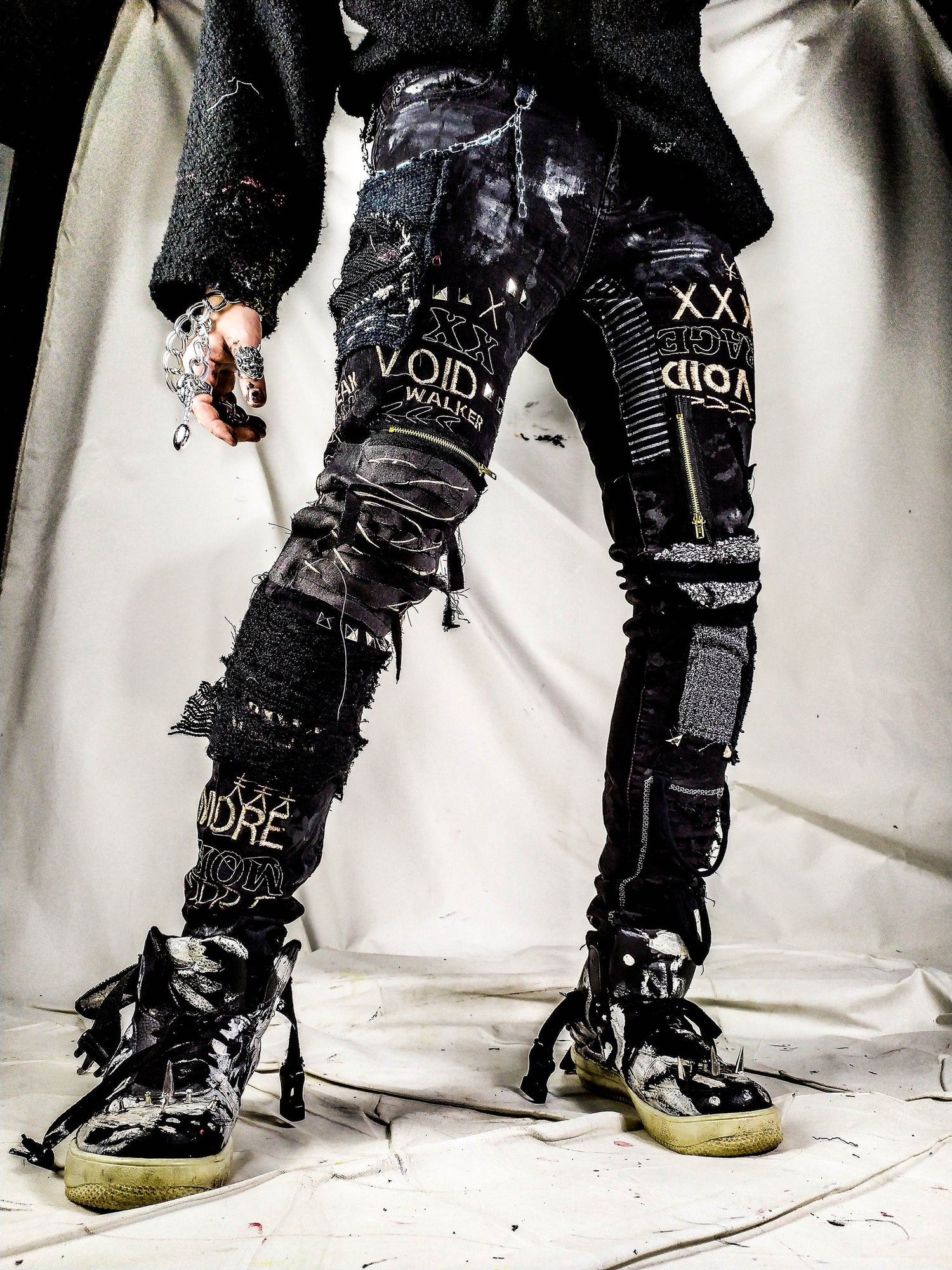 Model showing a black skinny pair with various abstract embroideries and words such as void walker and more riot with abstract symbols, black, white and grey patchwork, zippers, studs, splashes of gray and black paint and a chain attached to pocket.