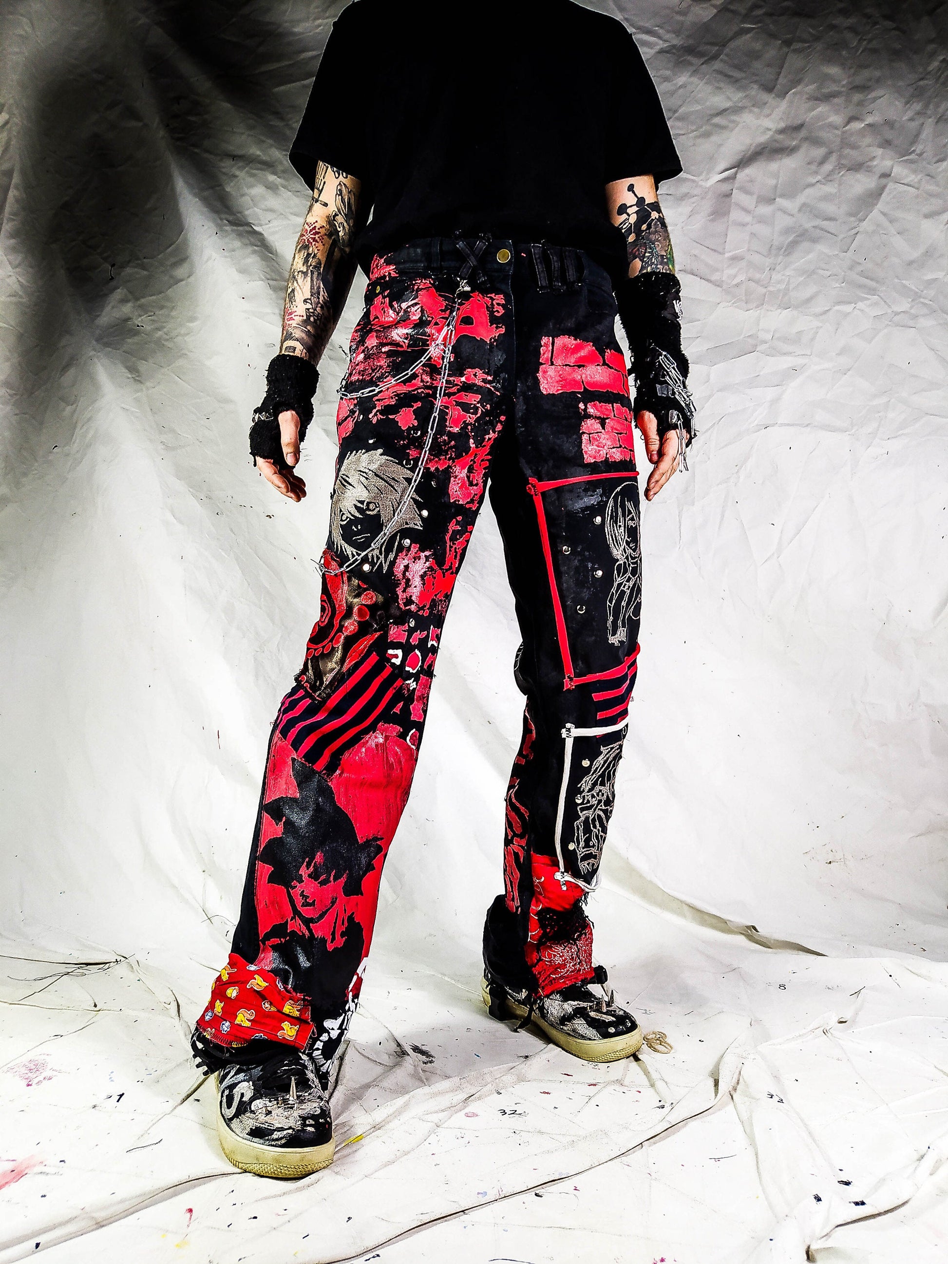 Model showing a pair of pants containing white embroideries with animes and a painted naruto, along with red patchwork, rivets, abstract paint, chains, the overall color scheme being black and red with a bit of white.