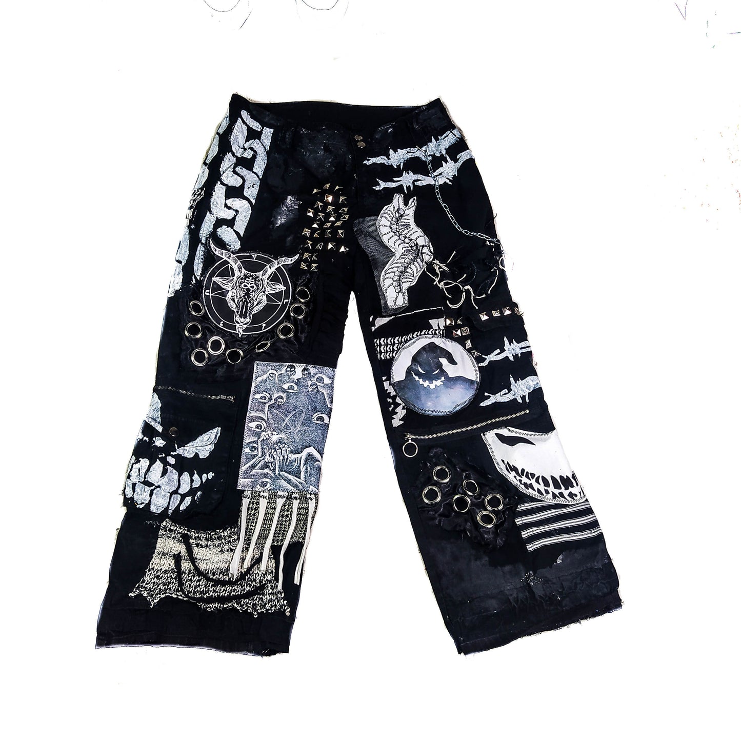 Flat laid black pair of baggy pants containing satanic, dark patches with a ghost, evil smiling faces, a goat with horns, a man screaming and eyes watching him, along with laces on patches, zippers, eyelets, a patch with messy threads, chain etc.