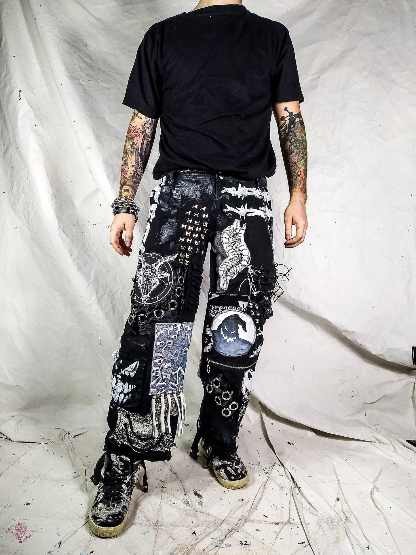 Model showing a black pair of baggy pants with satanic, dark patches with a ghost, evil smiling faces, a goat with horns, a man screaming and eyes watching him, along with laces on patches, zippers, eyelets, a patch with messy threads, chain etc.