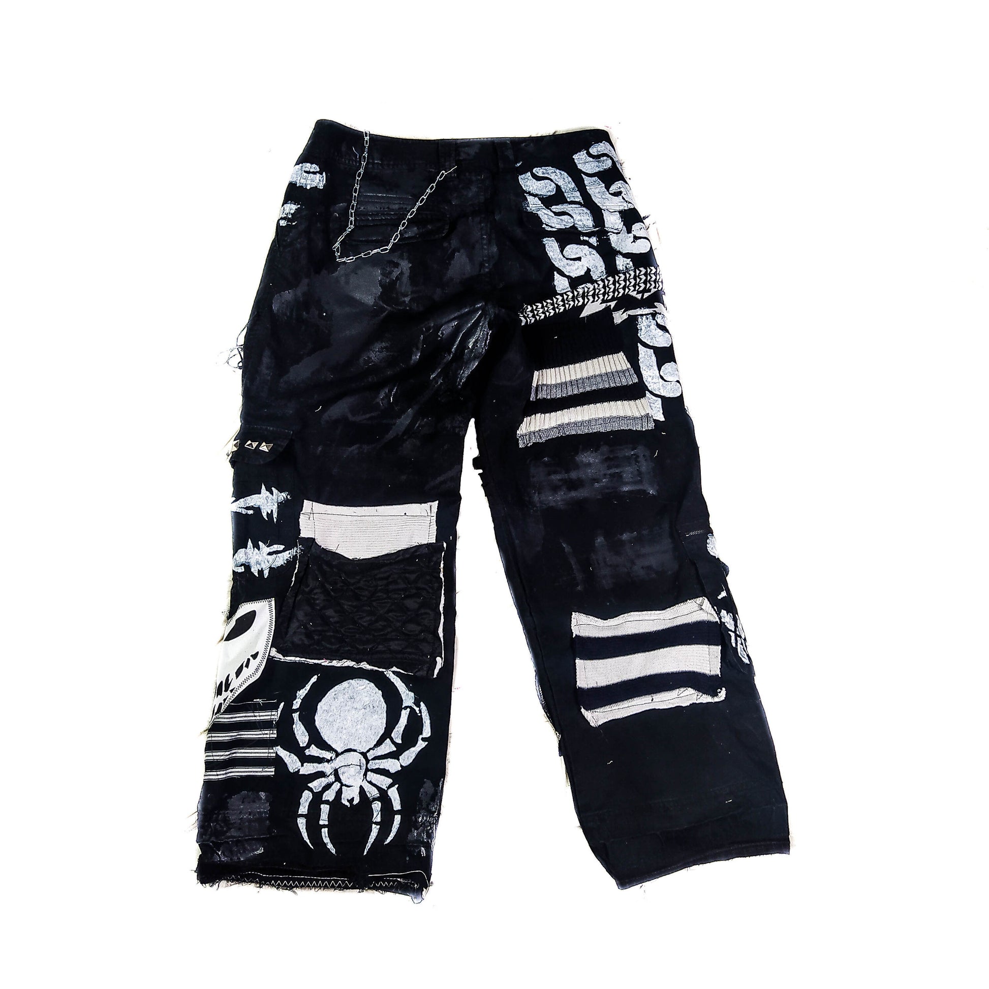 Flat laid pair of black baggy pants containing satanic, dark patches with a ghost, evil smiling faces, a goat with horns, a man screaming and eyes watching him, along with laces on patches, zippers, eyelets, a patch with messy threads, chain etc.