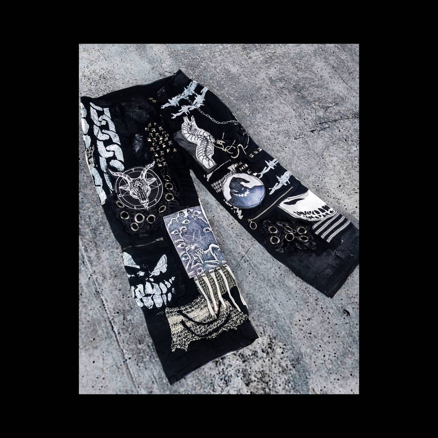 Flat laid black pair of baggy pants containing satanic, dark patches with a ghost, evil smiling faces, a goat with horns, a man screaming and eyes watching him, along with laces on patches, zippers, eyelets, a patch with messy threads, chain etc.