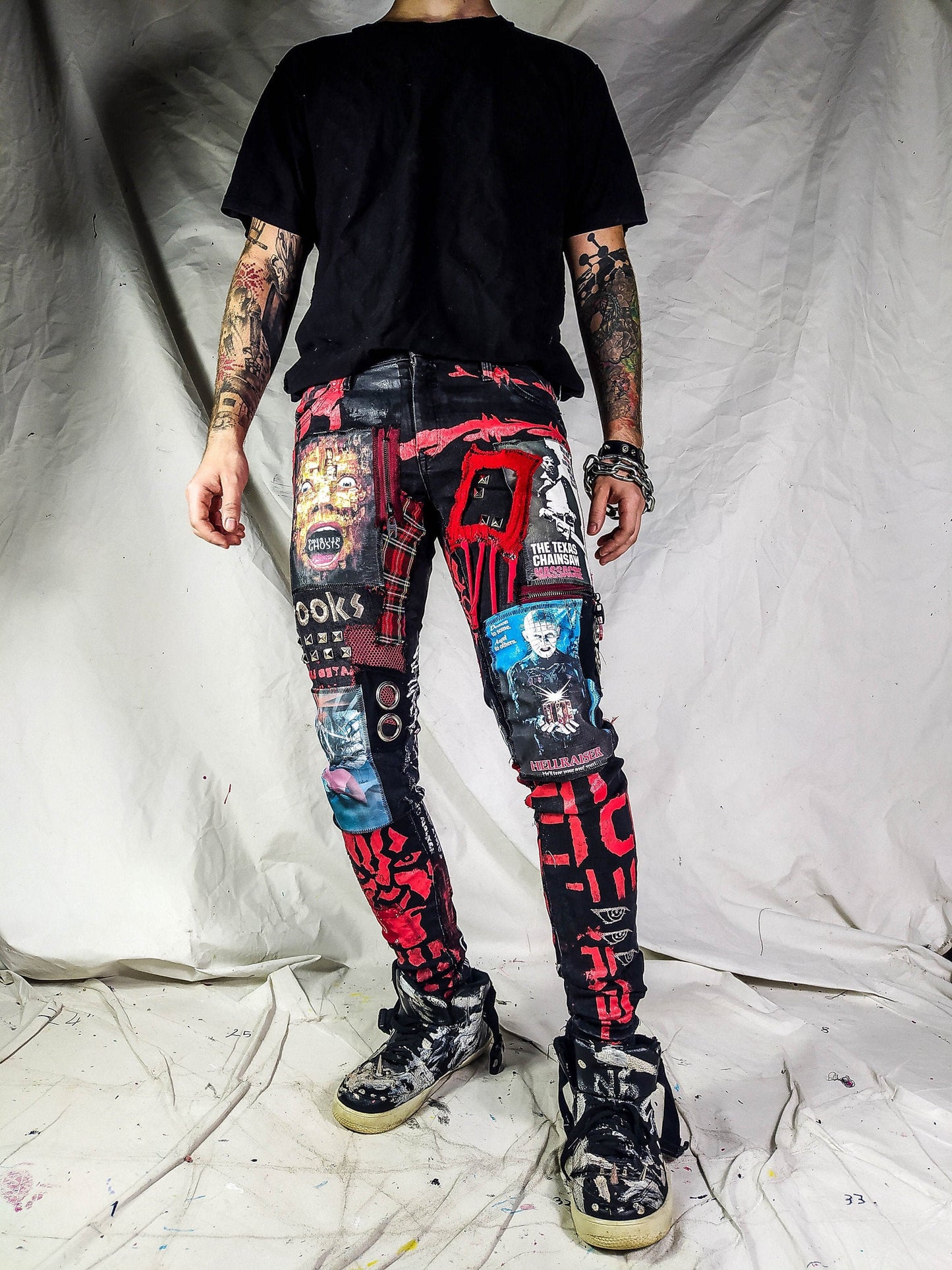 Model showing a skinny custom made black pair with prints containing various horror movies, red and black patchwork, abstract painting, studs, red laces hanging down, chain and eyelets.