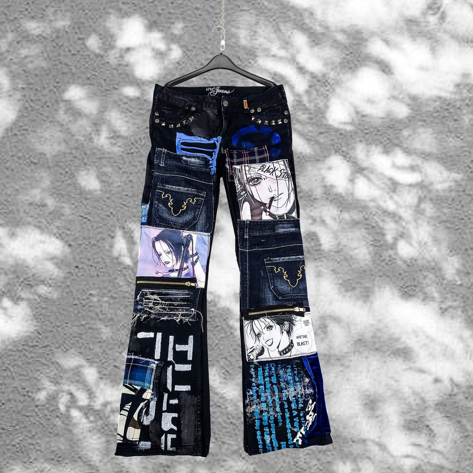 Flat laid pair of black pants with nana anime prints, pockets sewn on the pants, zippers, abstract patchwork and painting, color scheme is black and blue.