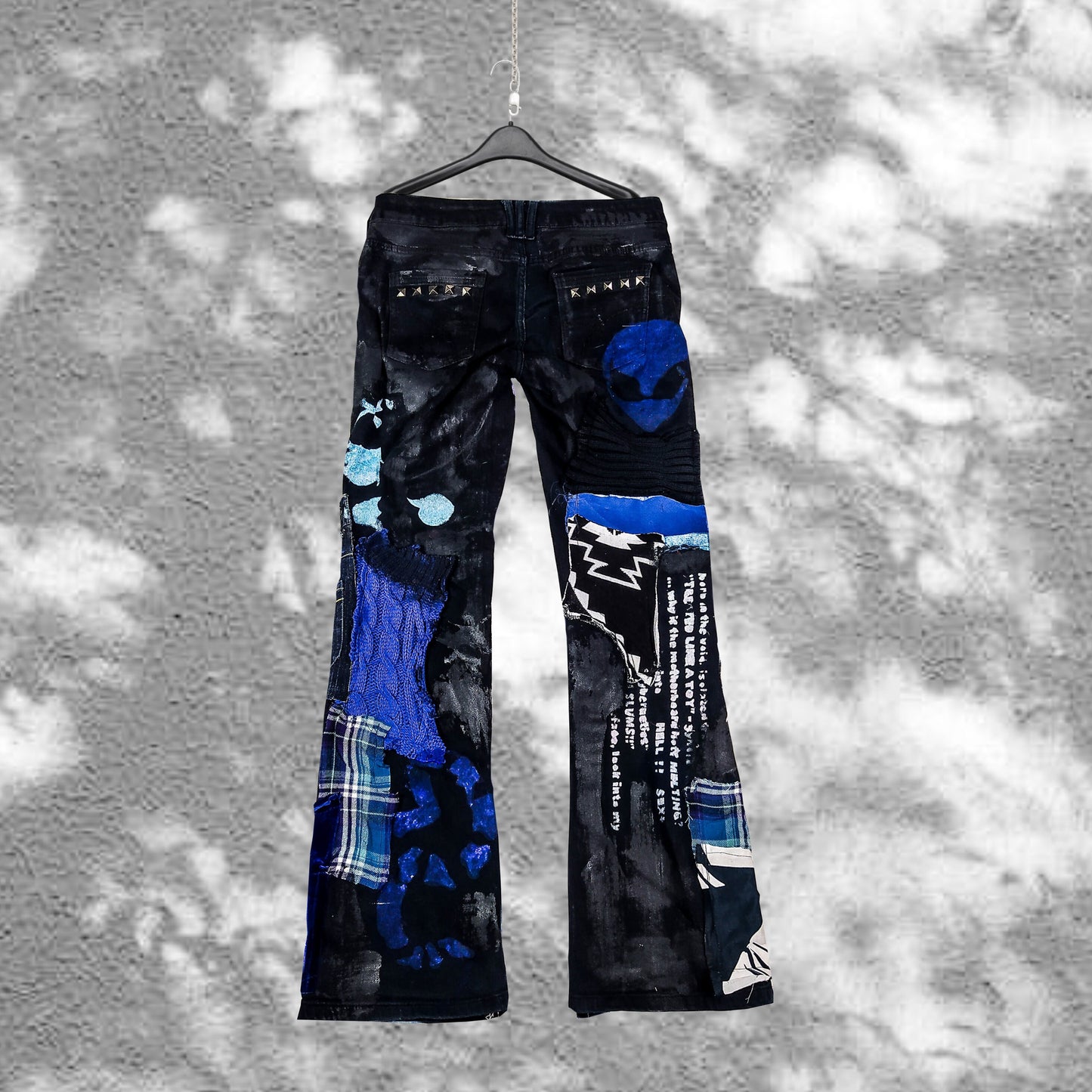 Flat laid pair of black pants with nana anime prints, pockets sewn on the pants, zippers, abstract patchwork and painting, color scheme is black and blue.