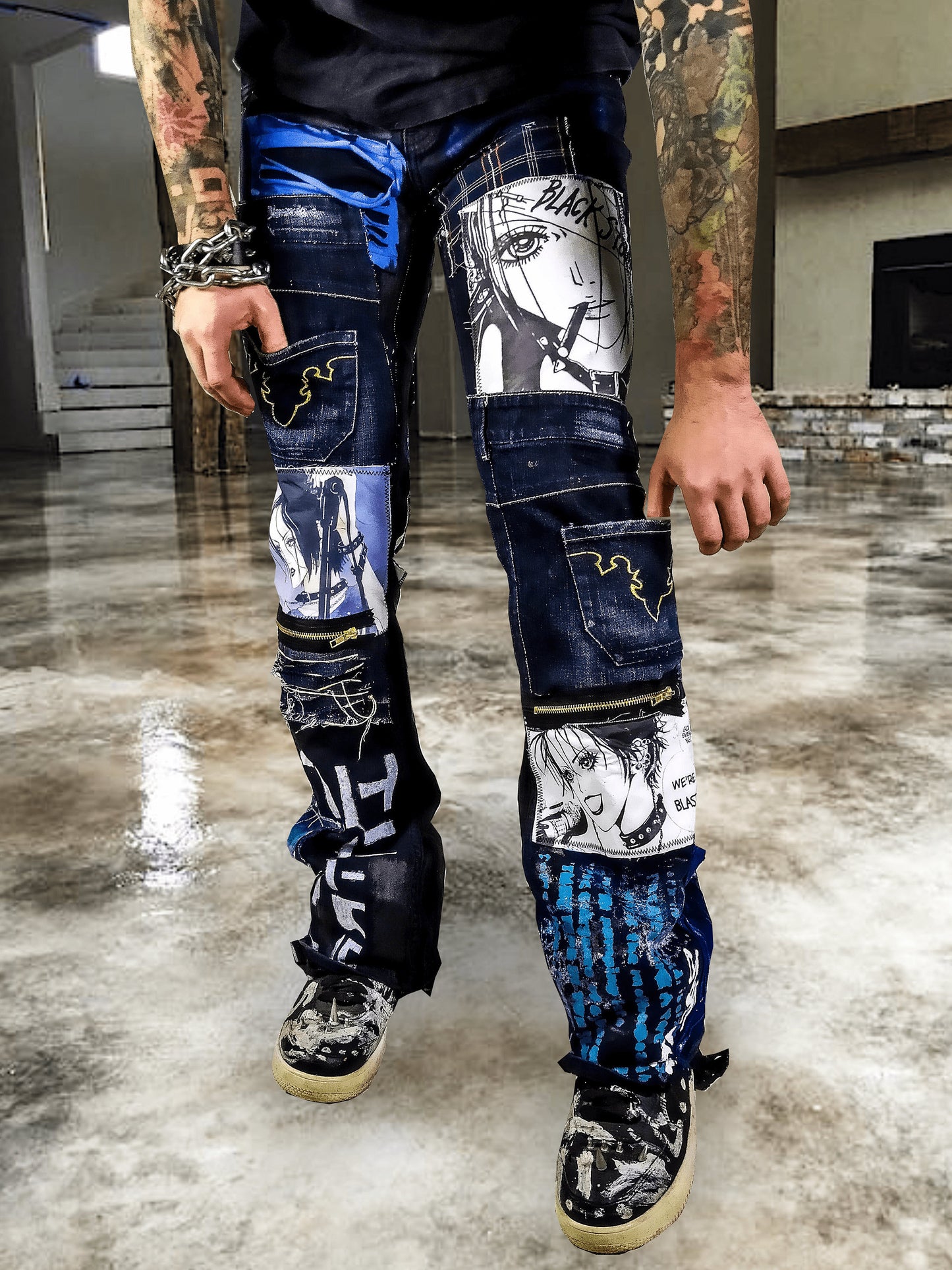 Model showing a black pair of pants with nana anime prints, pockets sewn on the pants, zippers, abstract patchwork and painting, color scheme is black and blue.