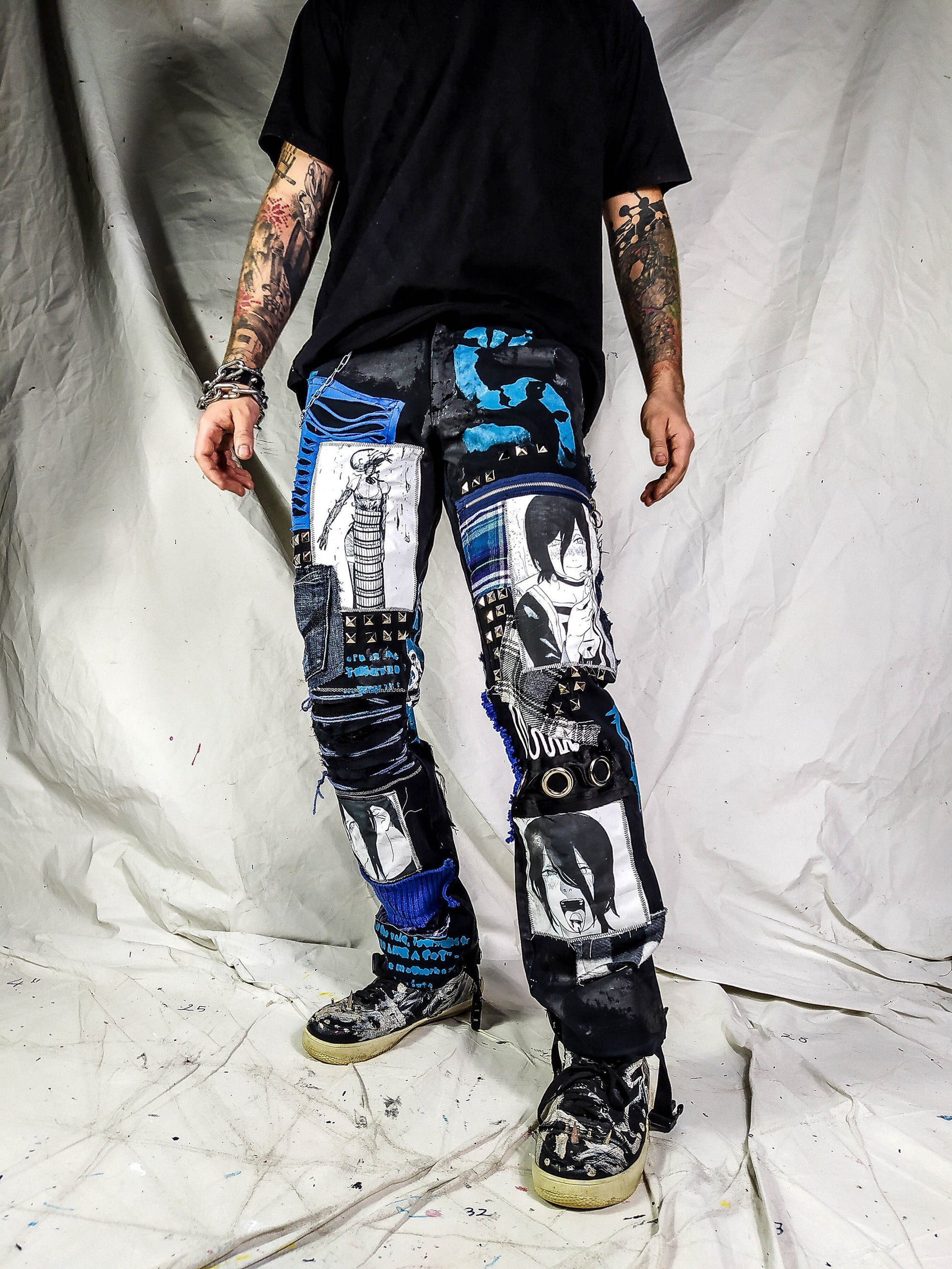 Model showing the black pants with anime prints, colorful patchwork in multiple blue shades, ripped patches, zipper, eyelets, studs and abstract painting with a blue snake on the front.