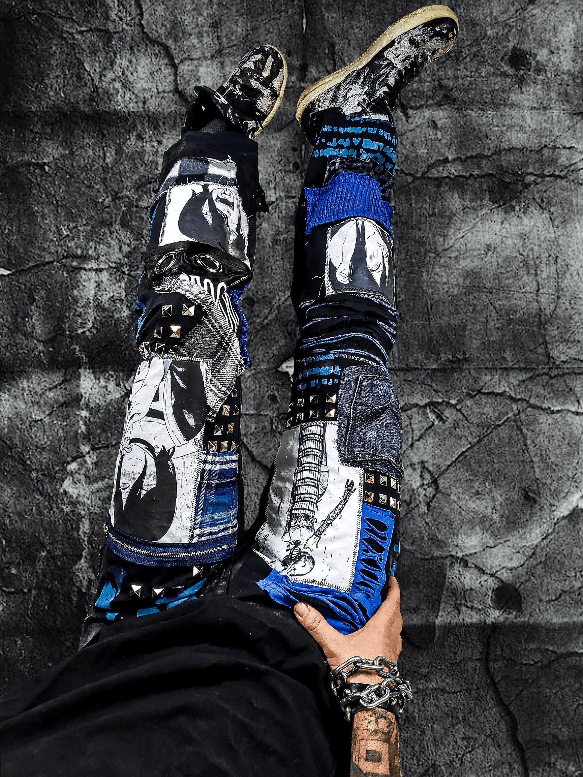 Model showing the black pants with anime prints, colorful patchwork in multiple blue shades, ripped patches, zipper, eyelets, studs and abstract painting with a blue snake on the front.