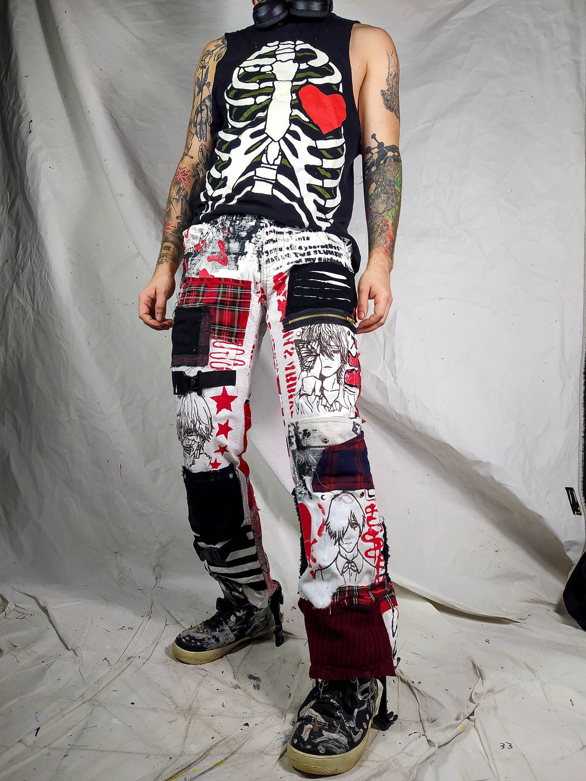 Model showing a pair of white pair of pants containing various anime embroideries with black and red patches, abstract black and red paintings, tags, rivets, buckle and chain.