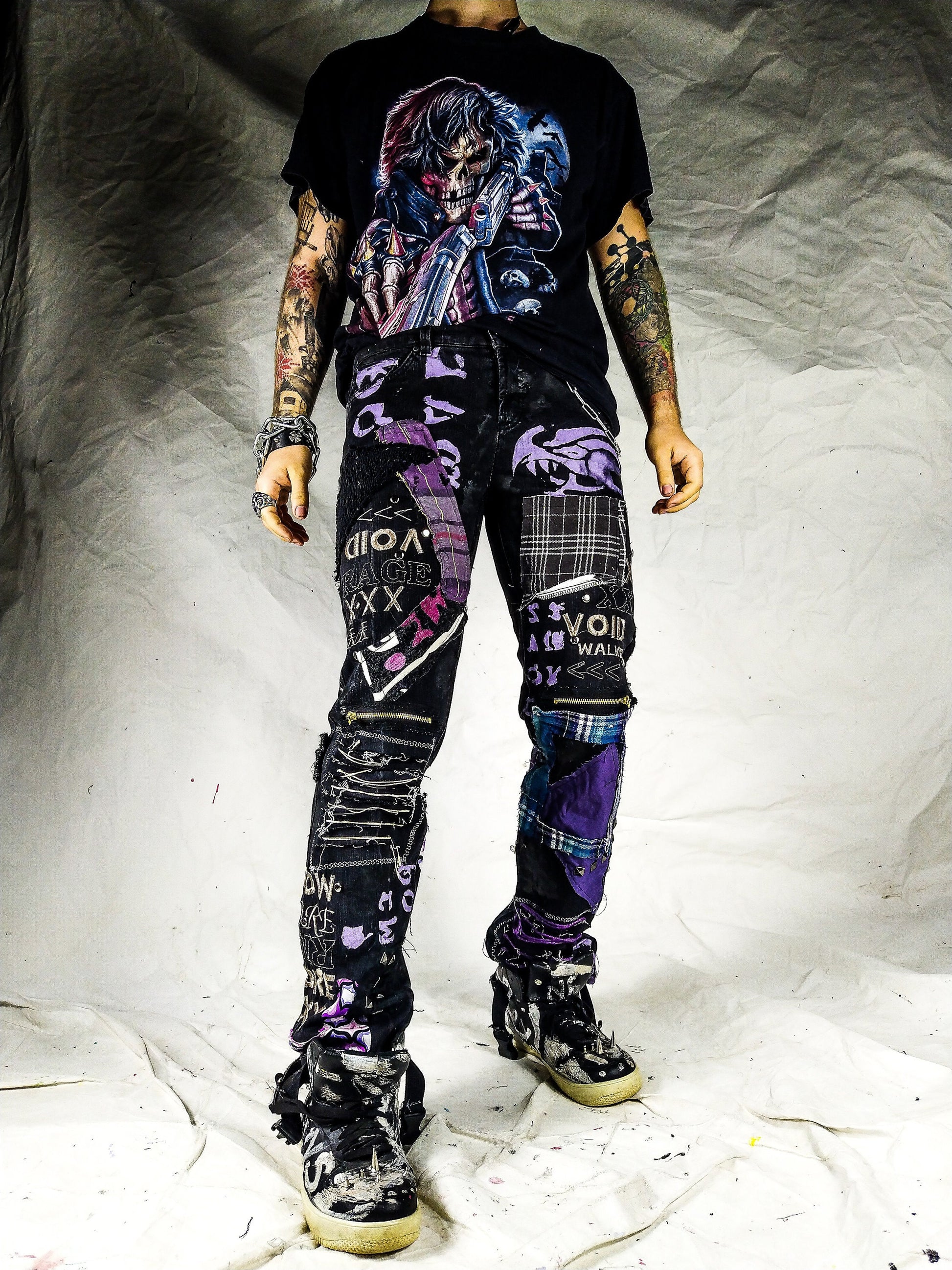 Model showing a black pair with blue and purple patchwork, embroidered words such as void walker and symbols, zippers, studs, ripped patches, chains, abstract painting.