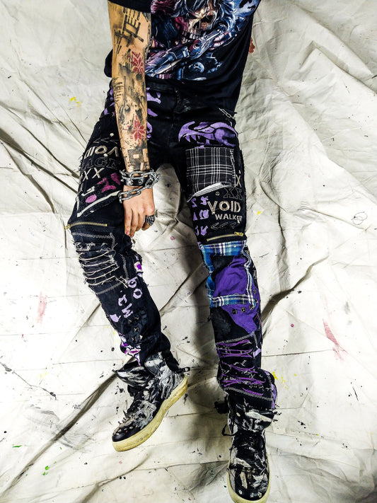 Model showing a black pair with blue and purple patchwork, embroidered words such as void walker and symbols, zippers, studs, ripped patches, chains, abstract painting.